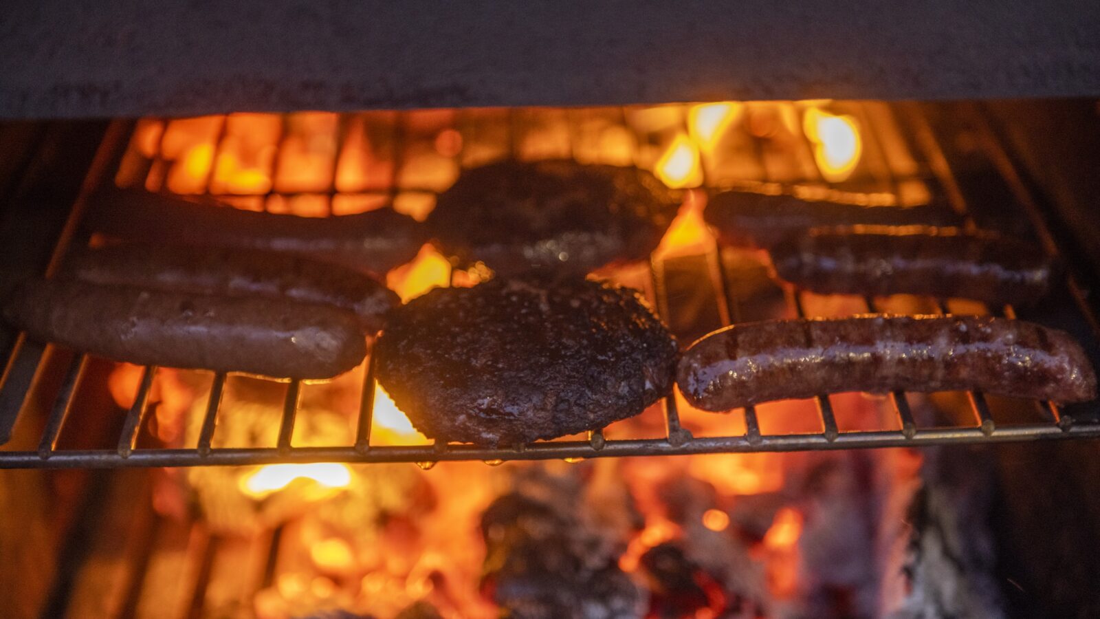 Close-up view of assorted meats grilling over an open flame. Two thick sausages and three round burger patties cook above hot coals and flickering flames, their sizzle echoing in the cozy borders of a tiny home's outdoor space, highlighting the intense heat and active cooking process.