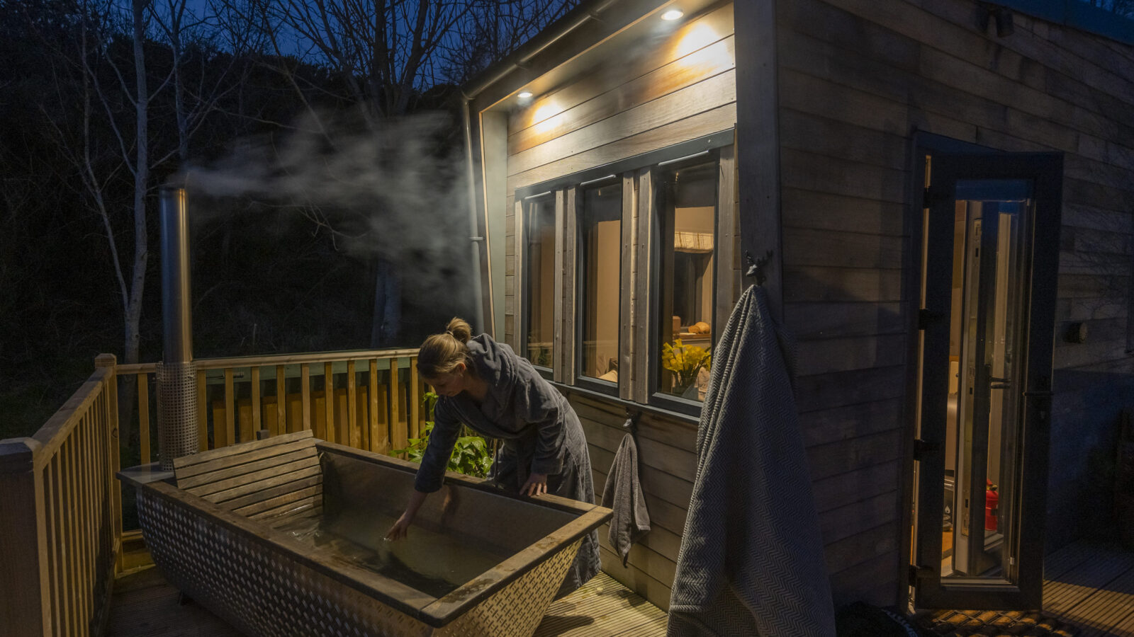 A person adjusts the water in an outdoor wooden hot tub next to a small modern tiny house at night. The cabin is illuminated by warm lights, and steam rises from the tub. The surrounding area is dark with bare trees, and a towel hangs on the cabin wall nearby.