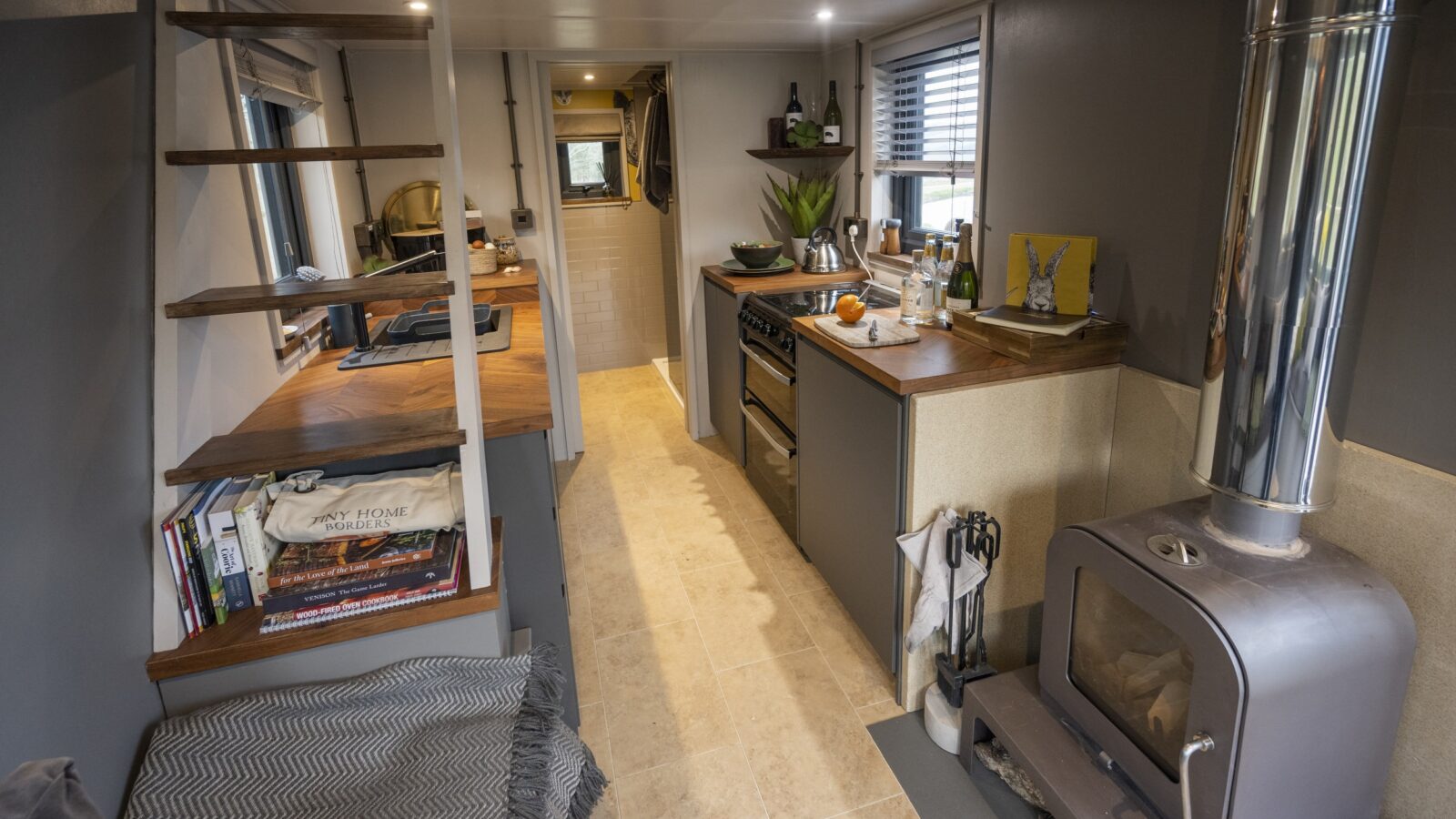 A cozy tiny home interior featuring a staircase on the left with open wooden shelves, a well-equipped kitchen with a stove and oven in the center, and a wood-burning stove on the right. The room has neutral tones, books, and a seating area with cushions and a blanket for those who love life within home borders.