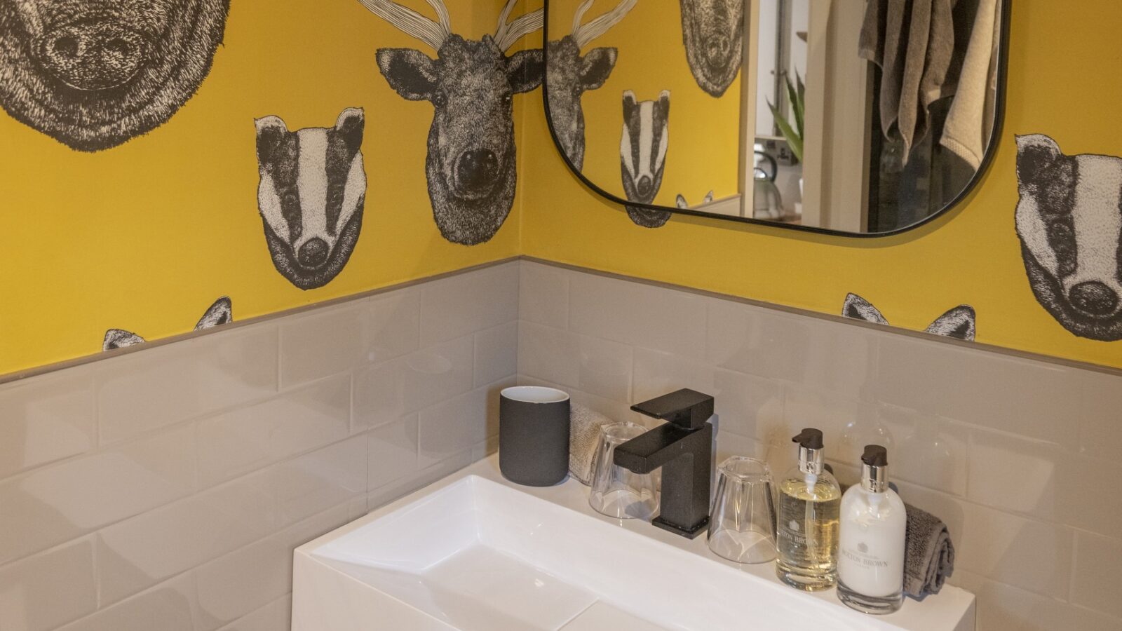 A modern bathroom features a sink with toiletries on a vanity against white tiled walls. The yellow wallpaper, adorned with black-and-white animal head illustrations—including a boar, deer, and rabbit—adds a unique touch. A square mirror is bordered elegantly and mounted on the wall, completing this cozy home space.