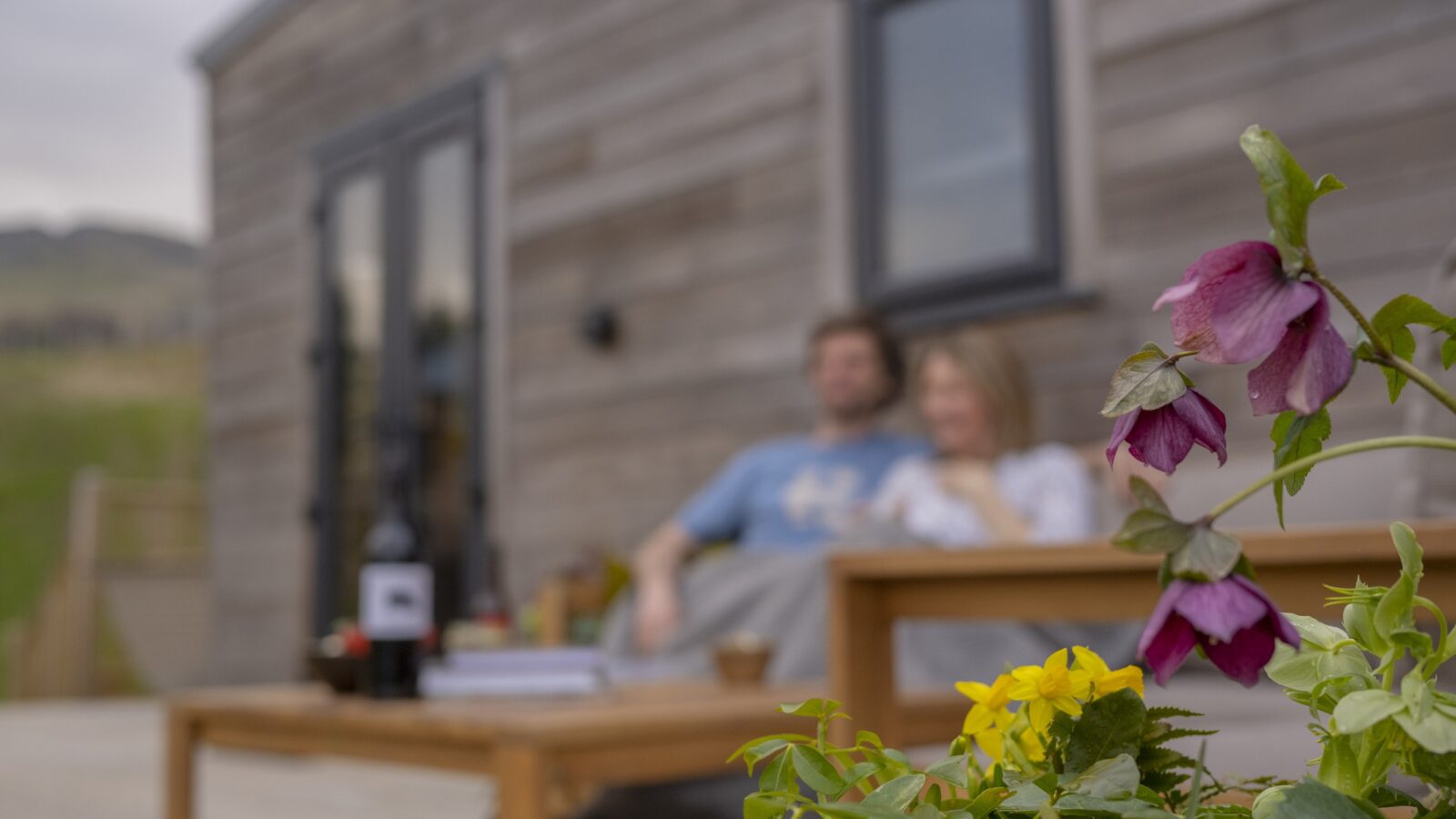 A couple sits on an outdoor patio, slightly out of focus, in front of a modern wooden tiny home. In the foreground, bright purple and yellow flowers are in sharp focus, contrasting with the blurred background. A coffee table with a bottle and books is nearby.