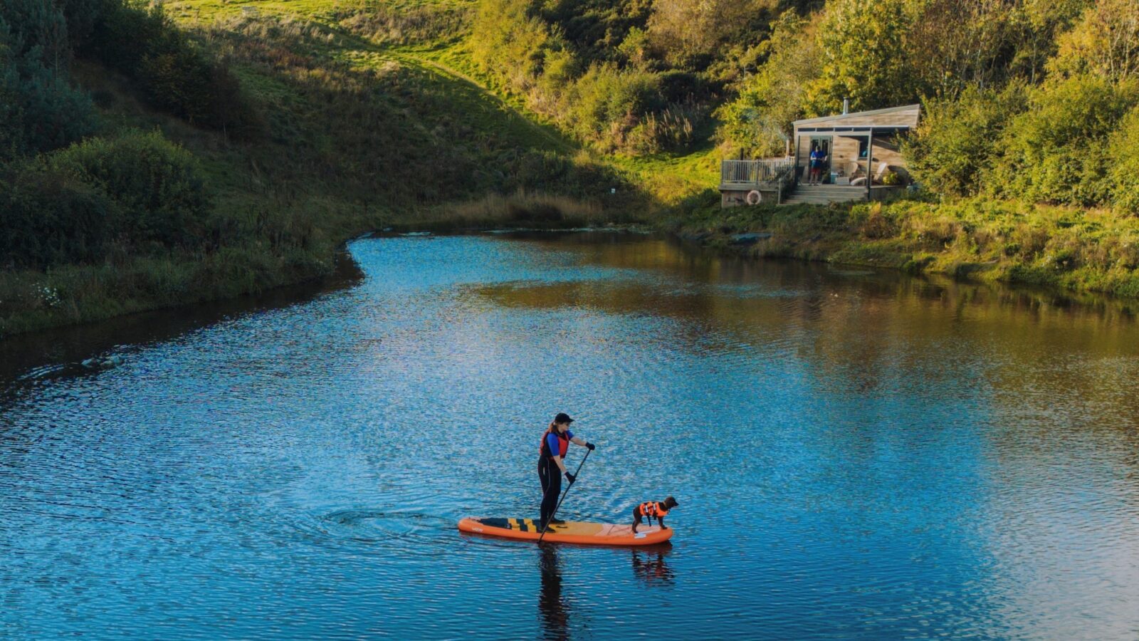 A person in a blue jacket and red life vest is paddleboarding on a calm river while holding a paddle. A small dog stands on the front of the paddleboard. Green hills with a tiny home nestled among them are visible in the background, adding charm as the clear sky borders the scene.