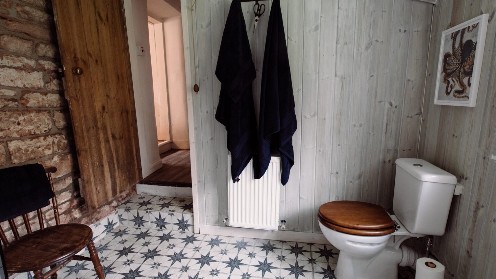 A rustic bathroom at Top Lodge features a tiled floor with star patterns, a toilet with a wooden seat, and a chair in the corner. A wooden door and exposed brick wall add charm. Towels hang on a hook above the radiator, and artwork adorns the wooden-paneled wall.