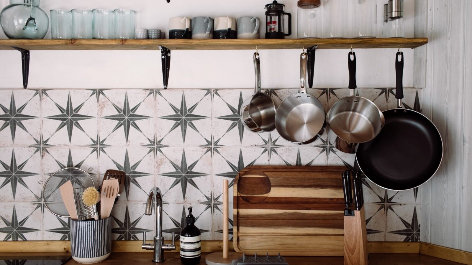 A top modern kitchen with a wooden countertop, sink, and utensils organized in a gray container. Above the sink, there are hanging pots and pans, a knife block, cutting boards, and a shelf with glasses, cups, and jars. The backsplash features star-patterned tiles that give it a cozy lodge feel.