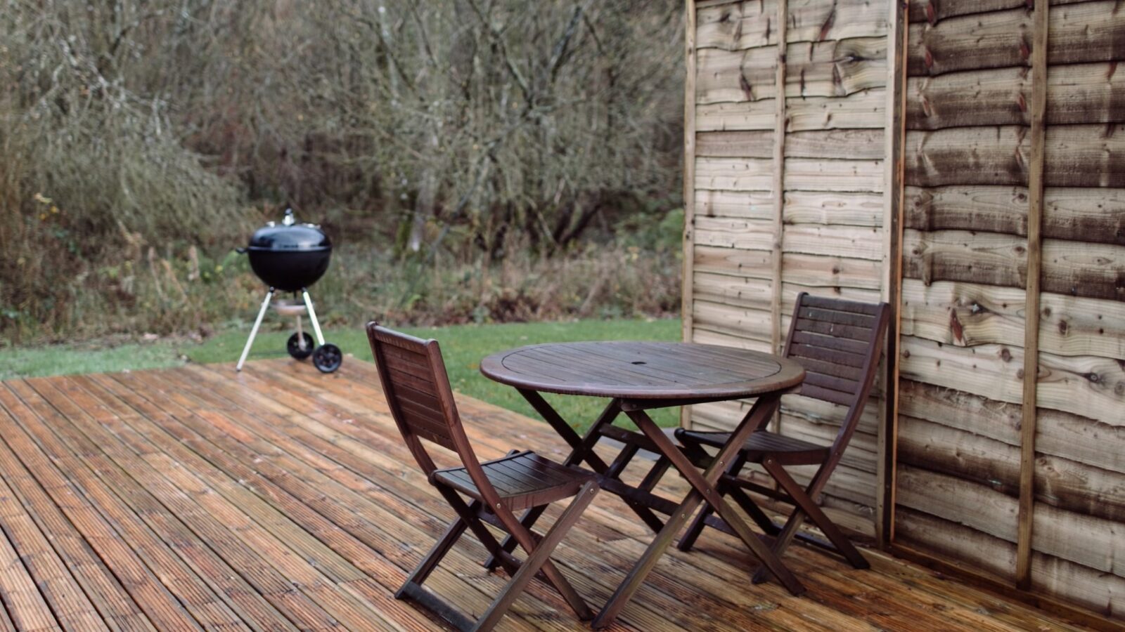 A wooden deck at the Top Lodge features a round table and two folding chairs. There is a wooden privacy screen on the right side. In the background, a charcoal grill stands on a grassy area next to dense, leafless trees, suggesting a late autumn or early winter setting.