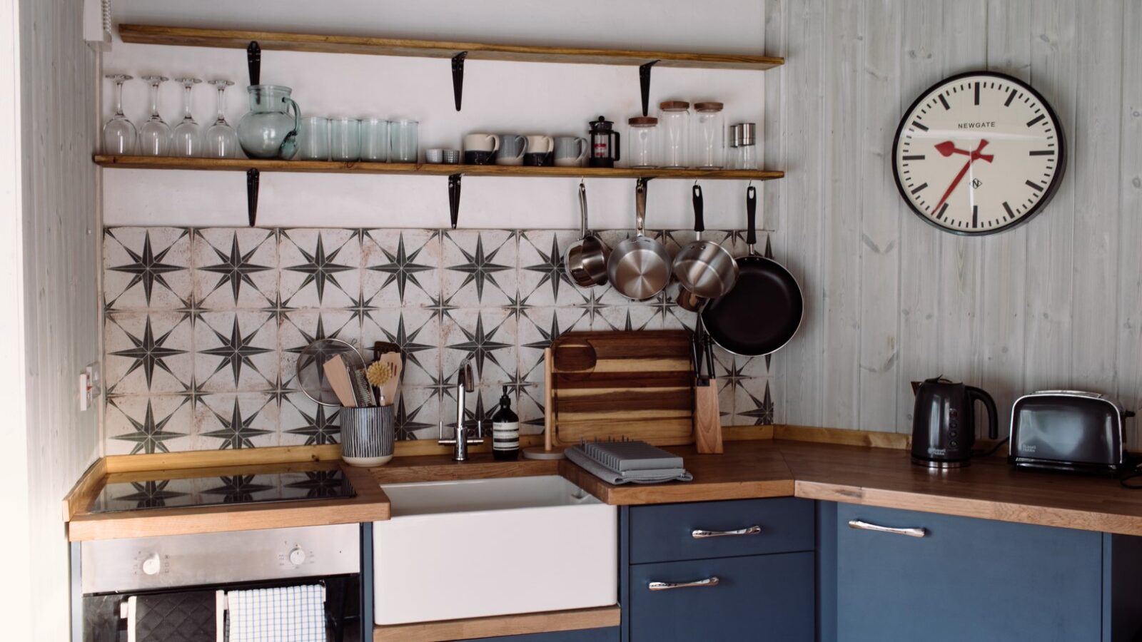 A cozy kitchen with blue cabinets and wooden countertops evokes a lodge vibe. Shelves above the sink hold glassware and jars. Pots and pans hang on hooks. A large clock on the wall shows 8:10. The backsplash features a black-and-white star pattern, and a toaster and kettle sit on the counter.