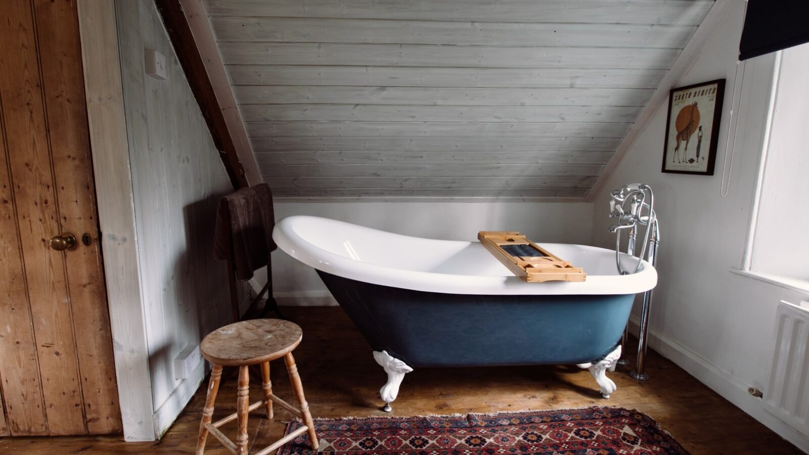 A cozy bathroom in Top Lodge with a freestanding bathtub under a sloped wooden ceiling. The tub, on a colorful patterned rug, features a wooden bath tray. A rustic stool is nearby, and an animal poster adorns the wall. The room is filled with natural light.