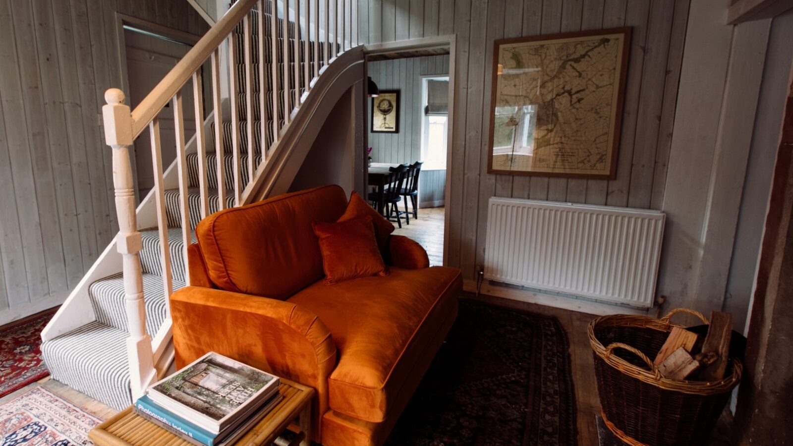 A cozy living room in Top Lodge features a bright orange sofa, a small wooden table with books, and a wicker basket in front of a white staircase. The light-colored wood walls boast a framed map and radiator. In the background, you can glimpse the inviting dining area.