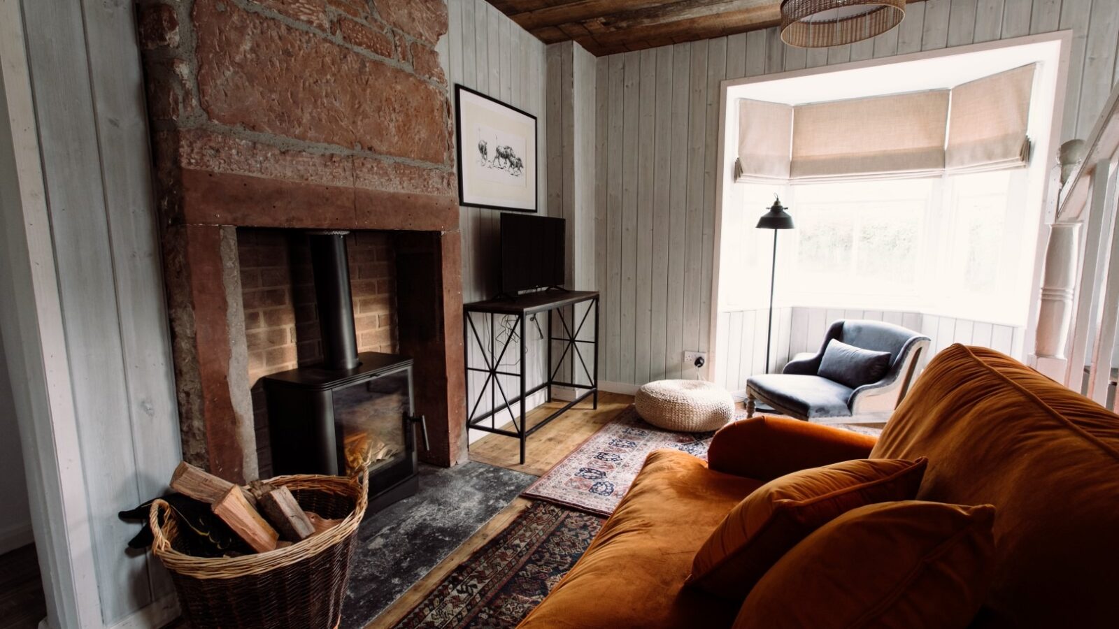 A cozy living room at Top Lodge features a wood-burning stove built into a brick fireplace, a wicker basket filled with firewood, an orange sofa with cushions, a black TV stand, an armchair, floor lamp, and a large window with light streaming through. The walls are wood-paneled.