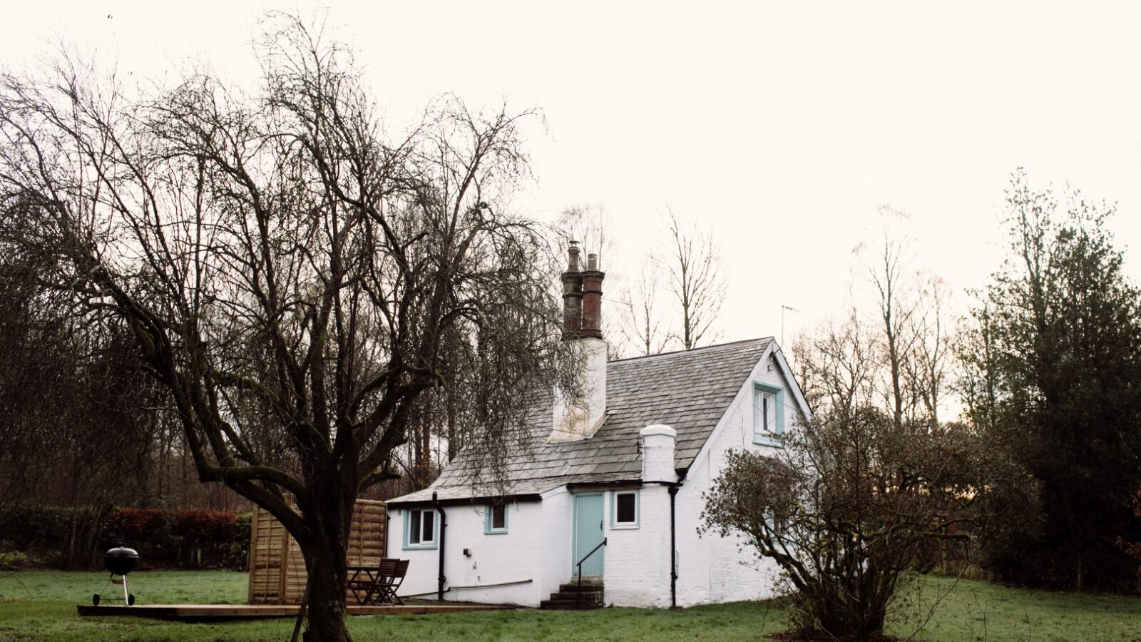 A small white cottage, known as Top Lodge, with a sloping roof and two chimneys stands in a grassy area. A large, leafless tree is to the left of the cottage, casting a shadow on the lawn. The sky above is overcast, and the surrounding trees are bare, suggesting a late autumn or winter season.