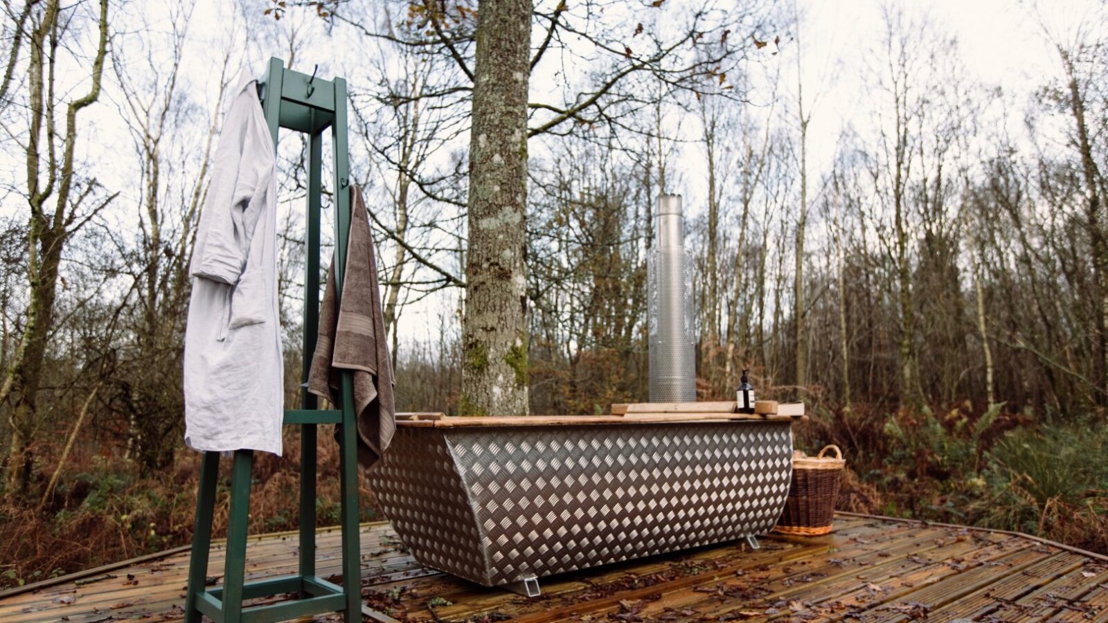 An outdoor bath setup on a wooden deck in a forested area provides the perfect retreat. The metal tub with diamond plate texture and chimney exudes rustic charm. At this top lodge, a green towel rack holds a white robe and brown towel, while a wicker basket and soap bottles sit nearby. Trees fill the background.