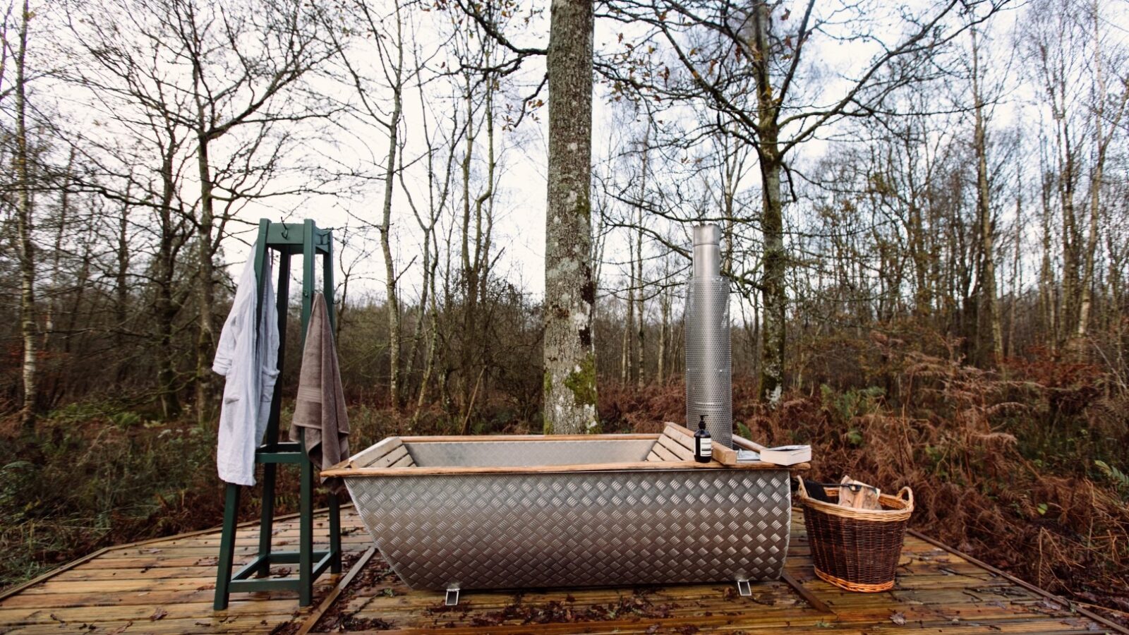 An outdoor bathtub is set on a wooden deck in a forested area, creating a top-notch lodge experience. Next to it, there's a towel rack with towels hanging and a wicker basket filled with firewood. Bare trees surround the deck, and the scene appears tranquil and secluded.