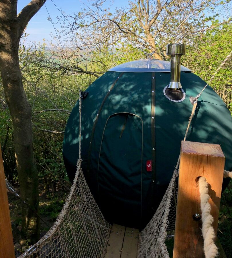 A round, green treehouse tent is suspended among the trees, connected by a rope bridge leading to a wooden platform. This idyllic setup on Pennard Hill features a tent with a small chimney and is situated in a lush forested area with fresh green foliage and branches visible in the background.