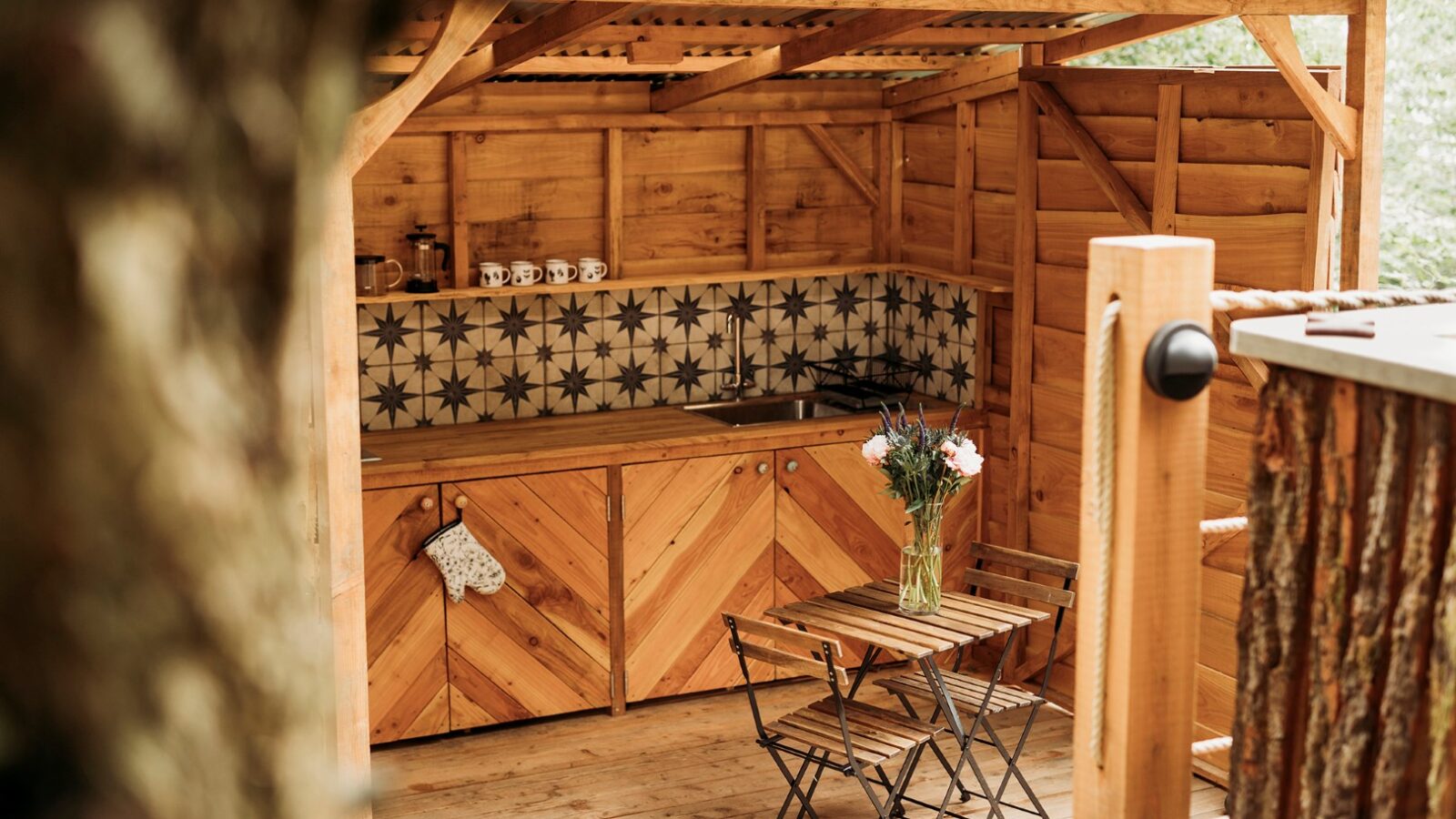 A rustic outdoor kitchen with wooden cabinets and a geometric-patterned tile backsplash. A small wooden table with two chairs and a vase of fresh flowers sit in front, all sheltered by a wooden pergola, reminiscent of the cozy charm found at Pennard Hill retreats.