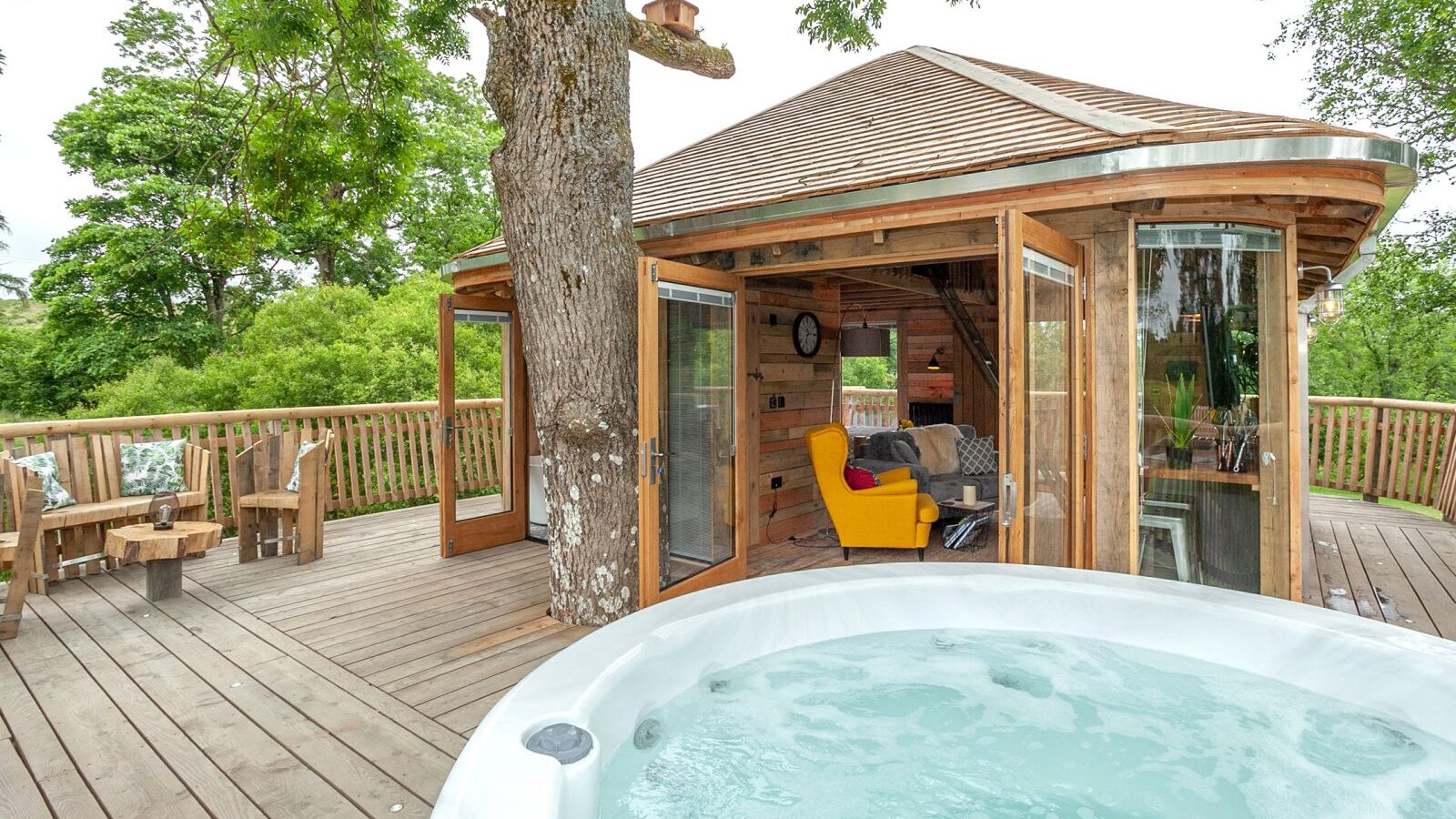 Discover the Trawscwm Treehouse, a unique accommodation featuring open glass doors, a wooden deck, a dining area, and a hot tub surrounded by lush greenery. Enjoy an unforgettable treehouse stay immersed in nature's tranquility.