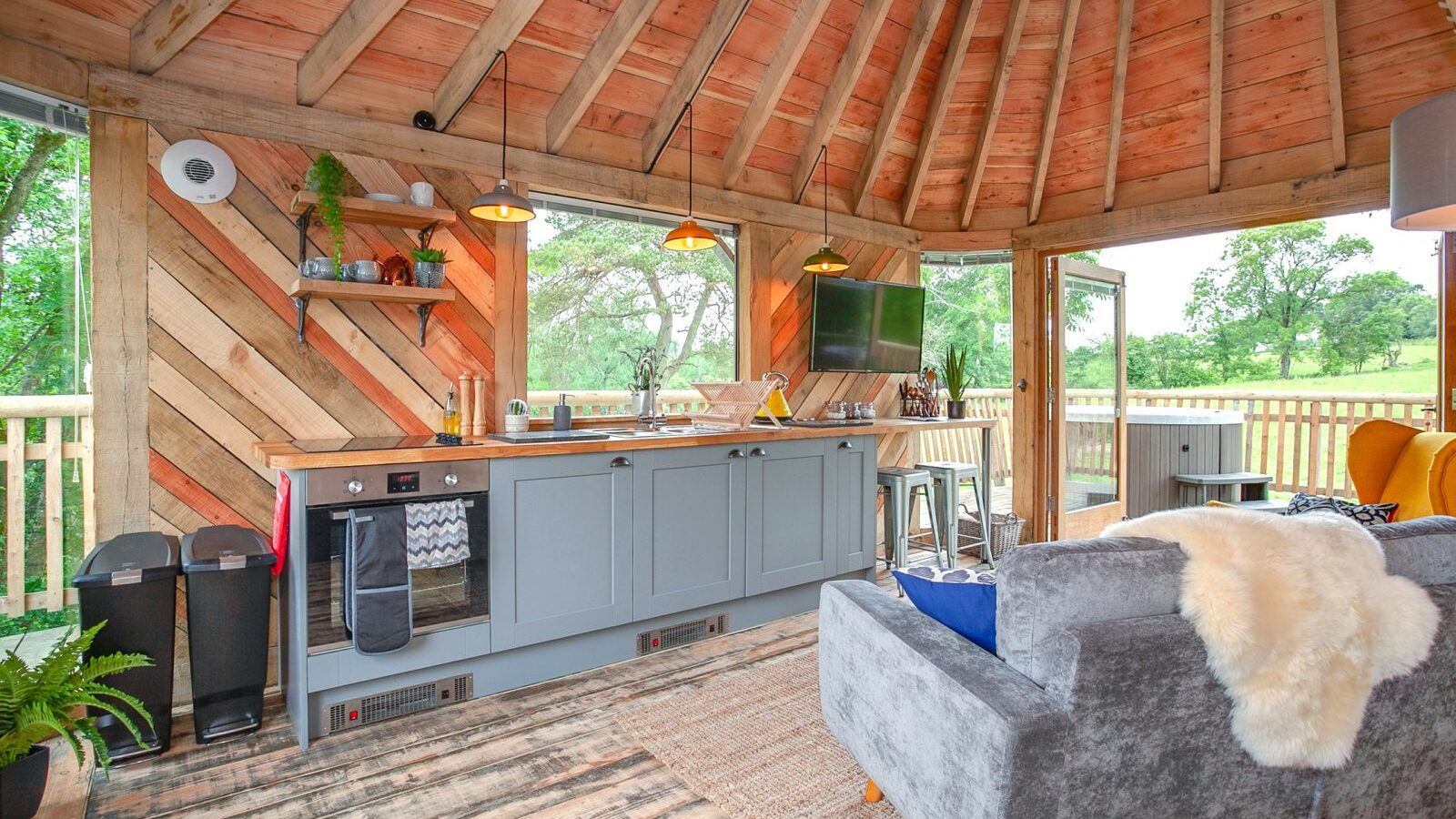 The Trawscwm Treehouse boasts a rustic open kitchen and living area with a wood beam ceiling, gray cabinets, modern appliances, and large windows that offer breathtaking views of the surrounding greenery.