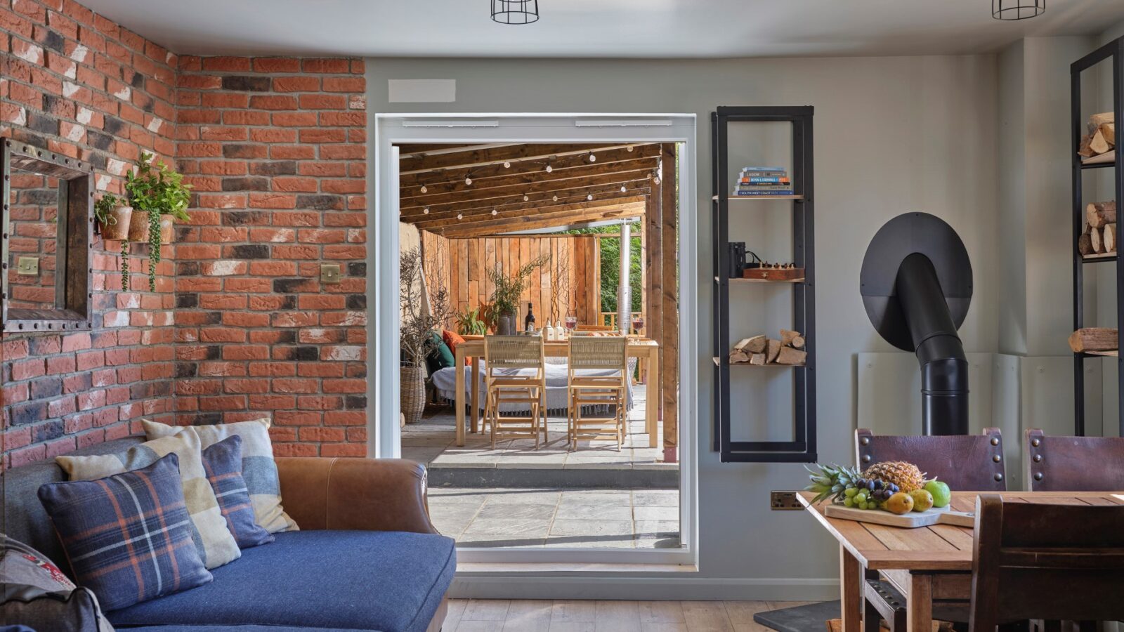 A cozy living room reminiscent of a Sleepy Owl retreat, featuring a blue sofa, a brick accent wall, and a wood stove. The room opens to a sunlit patio with dining set and wooden fencing. Decor includes a wall shelf with books and a table adorned with fruits, perfect for relaxed Devon afternoons.