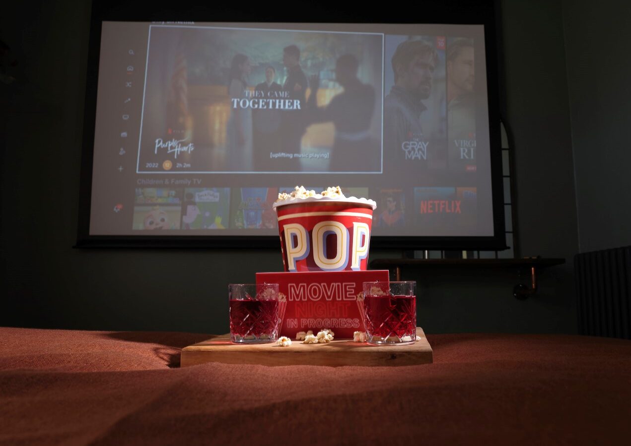 In a cozy setup reminiscent of a sleepy owl nestled in Devon, a home theater features a large screen displaying a movie streaming service. In the foreground, a box of popcorn and two glasses of red beverage rest on a wooden tray on the table.