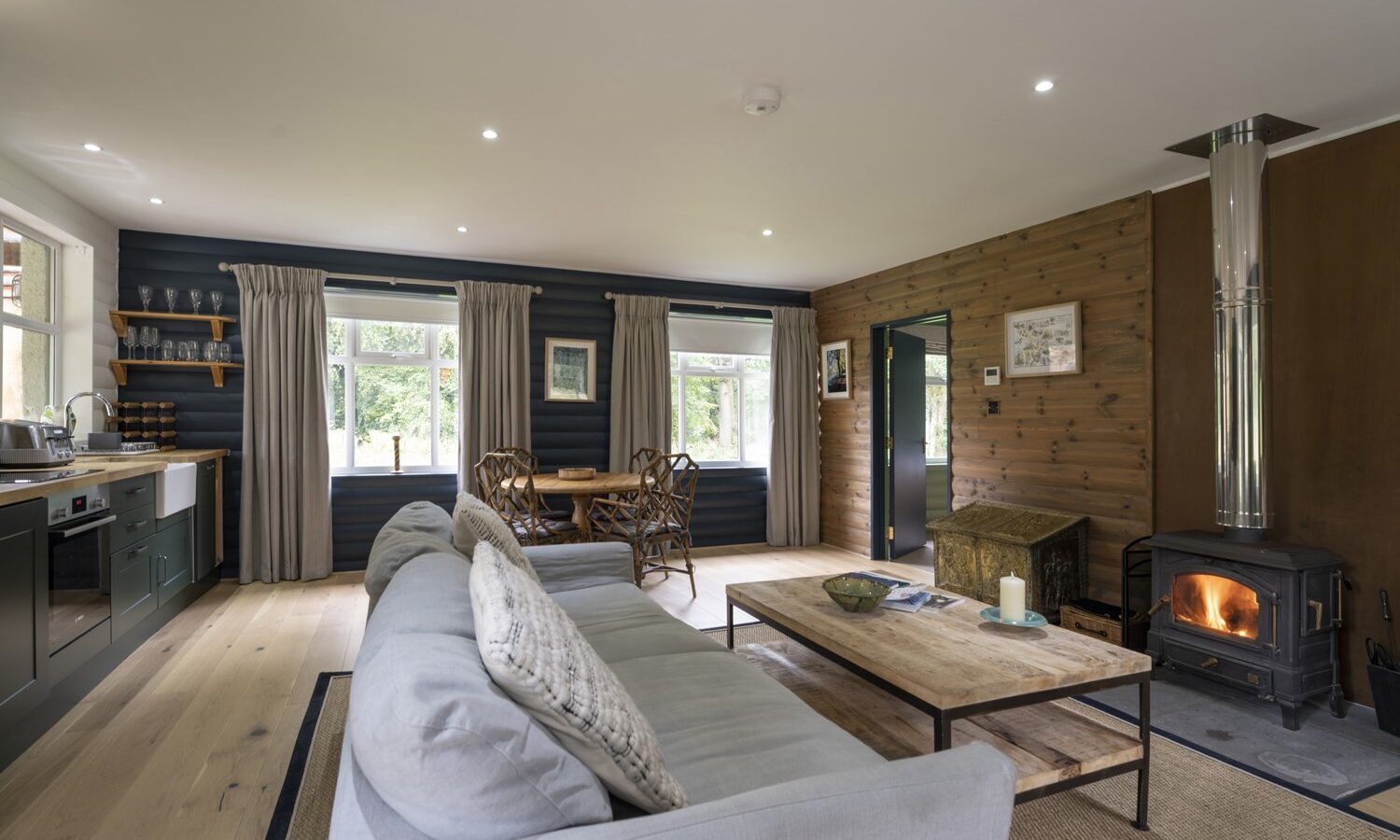 The cozy Keepers Bothy living room features a gray sofa, wooden coffee table, and a wood-burning stove. Large windows invite natural light into the space. Wooden walls complement an open kitchen with a dining area showcasing wicker chairs and a round table.