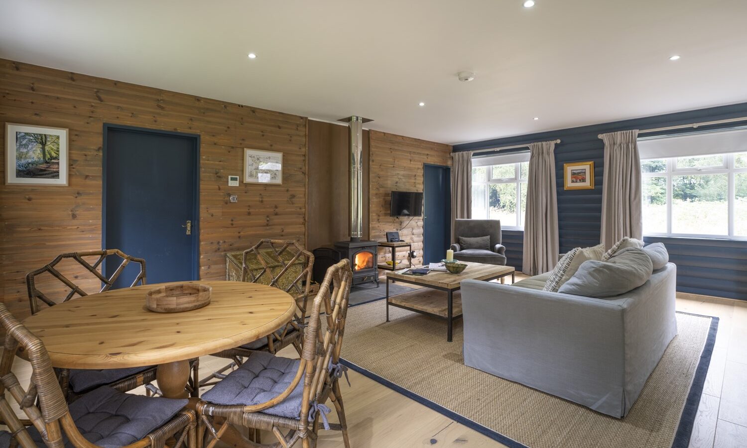 A cozy living room in Keepers Bothy, featuring wooden walls, a round dining table with wicker chairs, a gray sofa, a wood-burning stove, and a flatscreen TV. Large windows flood the space with natural light.