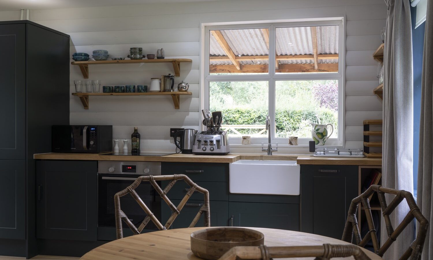 A cozy kitchen at Keepers Bothy features dark cabinets and wooden countertops. Shelves hold dishes and glasses, while a white farmhouse sink sits below the window. A coffee machine is ready on the counter, and a wooden table with wicker chairs invites travelers to unwind in this charming accommodation.