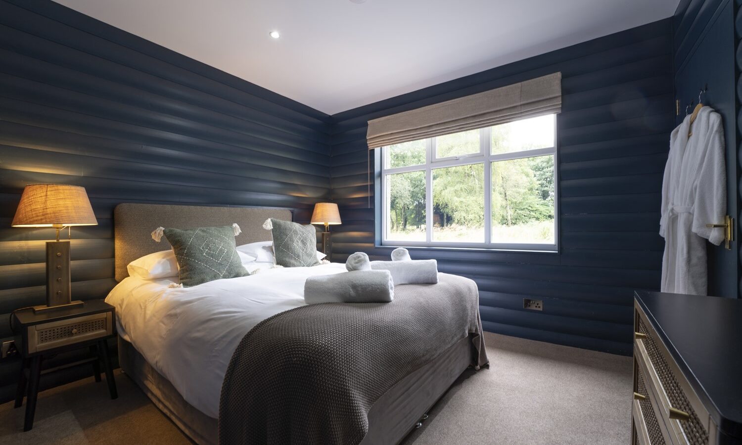 A cozy retreat bedroom with dark blue paneled walls and a large window. The bed, reminiscent of Keepers Bothy, features a gray headboard, white bedding, and green accent pillows. Two lamps are on nightstands beside the bed. Two white robes hang on the wall to the right—perfect accommodation charm.