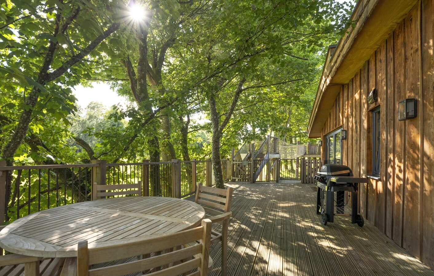 A wooden deck nestled in the canopy resembles a treehouse, surrounded by lush green trees. It features a round wooden table, chairs, and a barbecue grill. Sunlight filters through the leaves, casting dappled shadows and providing a serene escape for any visiting flycatcher.