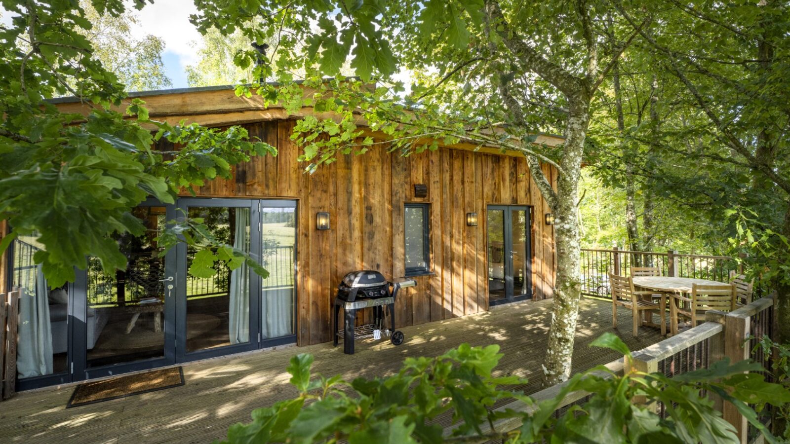 A wooden treehouse nestled in lush greenery, featuring large glass doors, a sundeck with a round table and chairs, and a barbecue grill. The leafy trees form a natural canopy, creating a serene and private setting where flycatchers flit about gracefully.