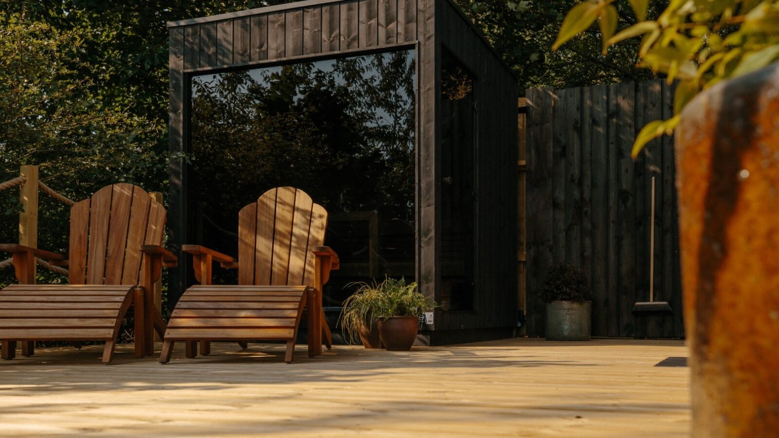 A cozy outdoor seating area with two wooden lounge chairs facing a small, modern, dark wood cabin with a large glass window. Sunlight filters through surrounding trees at Forcett Grange, casting shadows, and there are a few potted plants on the deck and near the cabin.