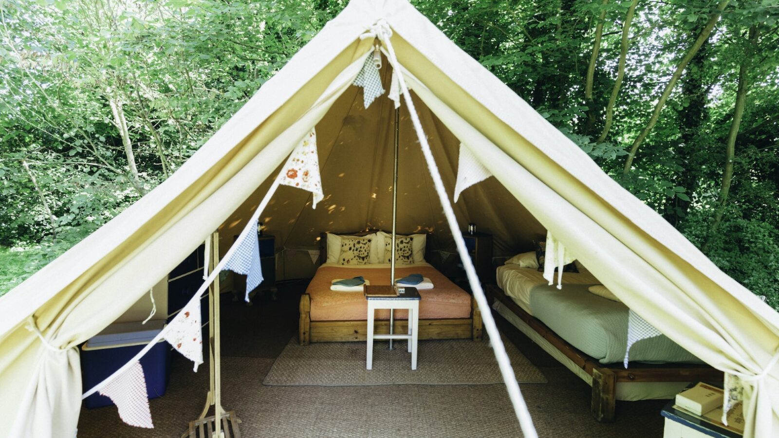 The brightly lit interior of a West Lexham bell tent features two cozy beds, a central table, and charming bunting decor, all nestled in a tranquil wooded area.