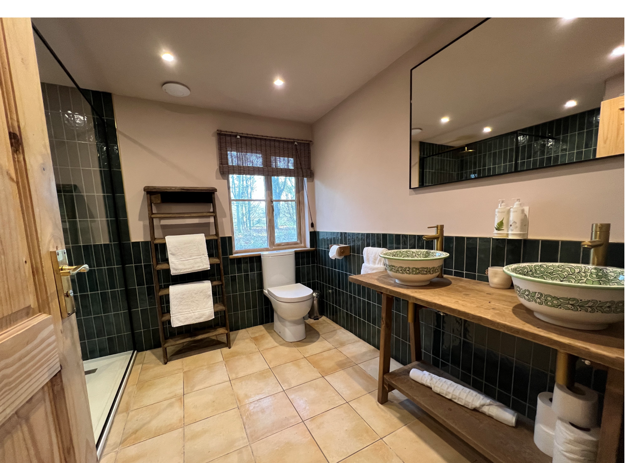 Discover a modern bathroom, perfect for a Secret Escape, featuring a sleek shower, double sinks, and wooden shelving. The tiled floor and walls complement the ambient light filtering through the window with blinds.