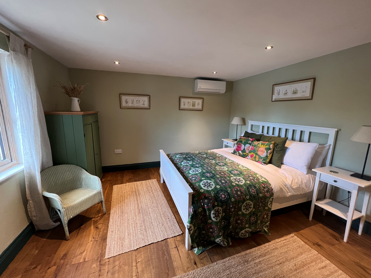 A cozy bedroom by Browning Bros features a double bed with a green quilt, two bedside tables, and a wicker chair. Framed art adorns the light green walls, creating a tranquil retreat perfect for secret escapes.