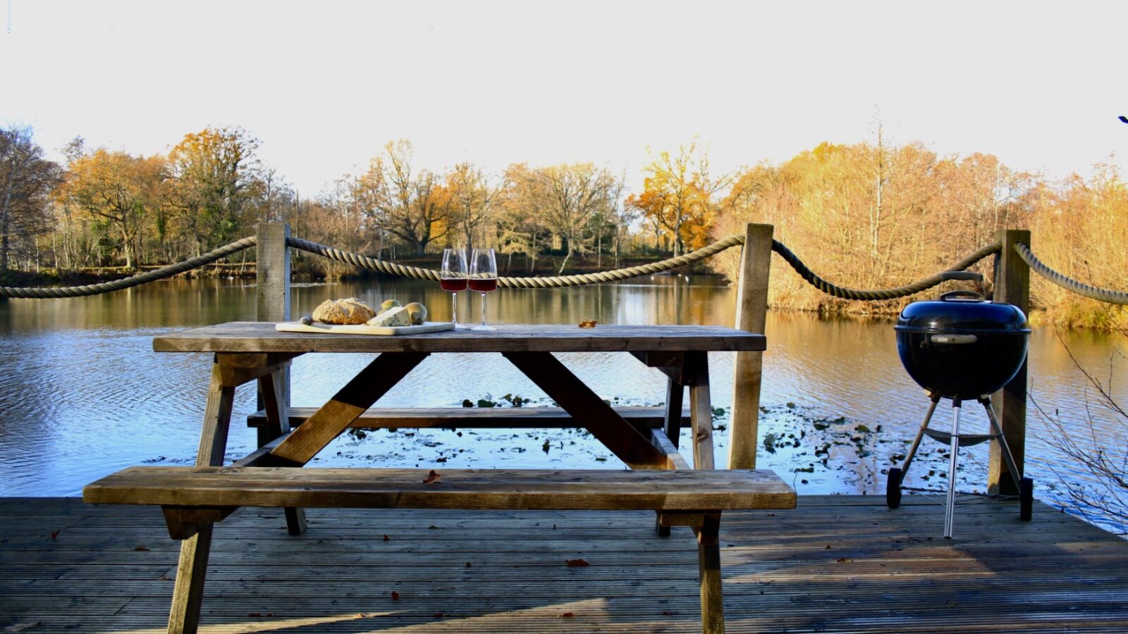 A wooden picnic table adorned with bread, cheese, and two glasses of wine is set by a lakeside under the golden hues of autumn trees, offering a scene reminiscent of Browning Bros' Secret Escapes.