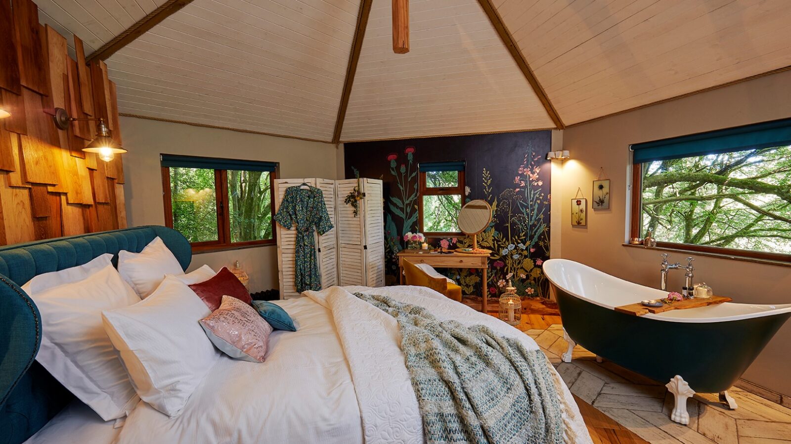 A cozy bedroom in the charming Ravendere Treehouse features a vaulted ceiling, a queen-sized bed with white linens, a teal headboard, and a patterned blanket. In the corner, there's a green freestanding bathtub next to a window. A room divider and rustic vanity are also present, with greenery visible outside the windows.