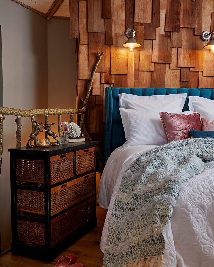 A cozy bedroom at Ravendere Treehouse features a bed with blue upholstery, white bedding, and pink and blue pillows. A woven blanket is draped on the bed. A rustic wooden nightstand holds a lamp, flowers, and decor items. An accent wall with wooden panels and two wall-mounted lights add warmth.