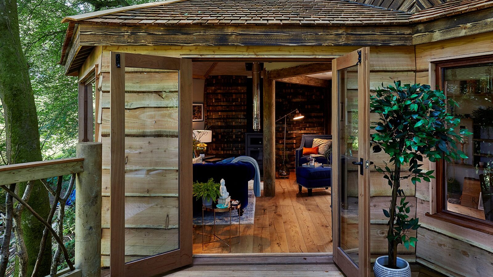 A rustic Ravendere Treehouse with open French doors leading to a cozy interior. The room features wooden walls and floors, a blue couch, a coffee table, bookshelves, and a floor lamp. Outside, there's a small porch with railing and a potted plant next to the door.