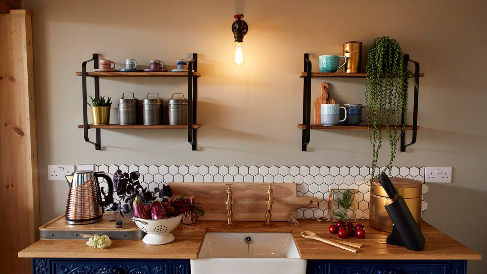 A cozy kitchen with wooden countertops and colorful decor, reminiscent of a Ravendere Treehouse nature retreat. Shelves above hold cups, spices, and a potted plant. The counter features a colander with vegetables, cooking utensils, and a knife set. A vintage-style bulb illuminates the hexagonal backsplash tiles.