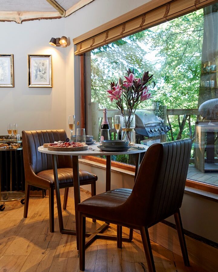 A cozy corner dining area in the Ravendere Treehouse features a small, round table with two brown leather chairs. On the table are wine glasses, a bottle of wine, a plate of food, and a vase with pink flowers. A large window offers a view of lush green trees outside.