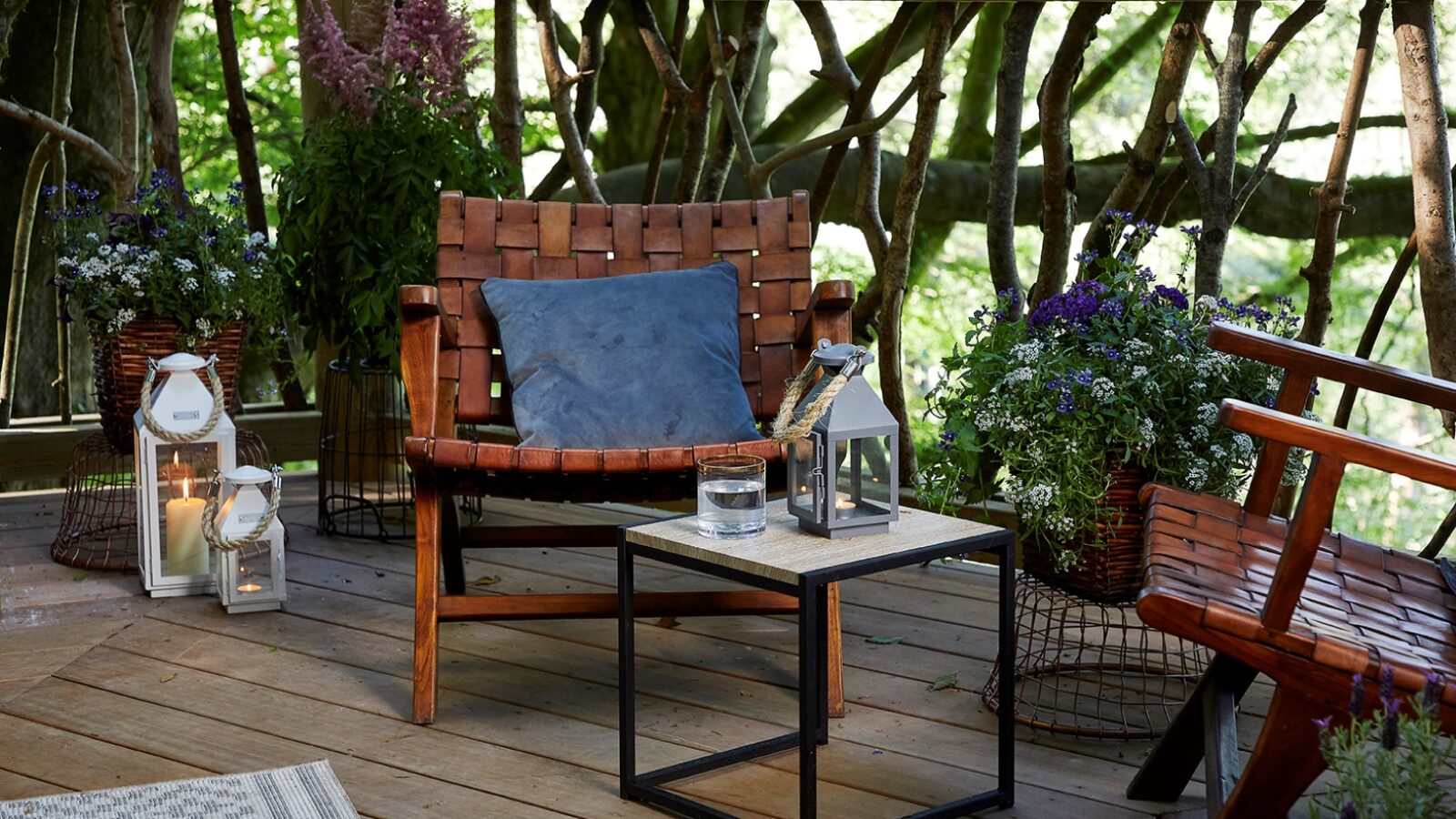 A cozy outdoor seating area on the wooden deck of Ravendere Treehouse features a woven leather chair, a blue cushion, and a small table holding a glass of water, two white lanterns, and a potted plant. Lush greenery and additional flower arrangements surround the tranquil space.