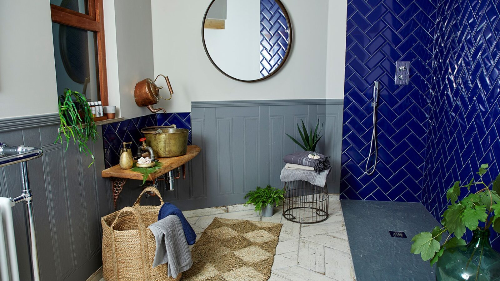 A modern bathroom in the Ravendere Treehouse features blue herringbone tile walls in the shower, a round mirror above a wooden sink countertop, copper fixtures, a woven basket with towels, and a small rug on a patterned tile floor. Plants add a touch of greenery to these luxury accommodations.