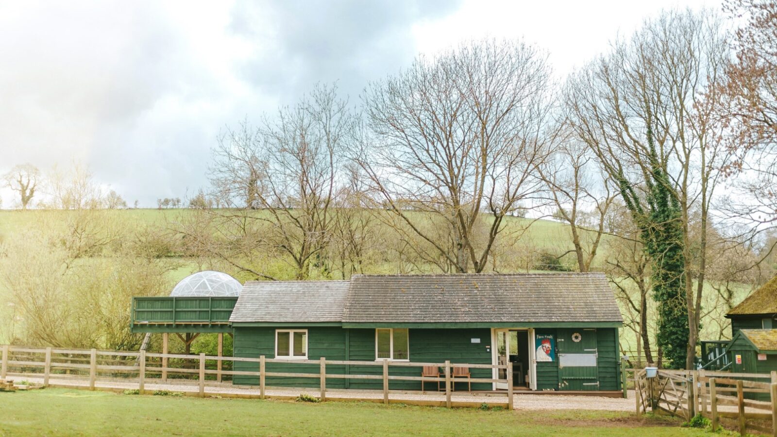 A small green cabin with a domed extension sits in a rural landscape, resembling a quaint lodge surrounded by bare trees and a wooden fence.