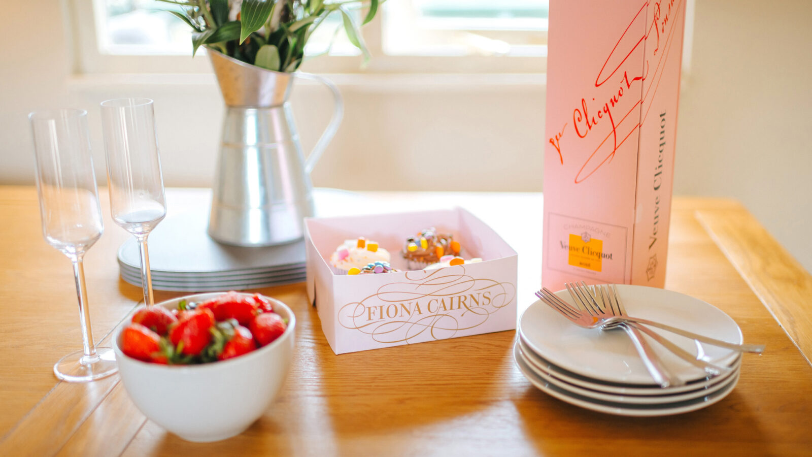 A table set at Chicken Shed Lodge with two empty champagne glasses, a metal jug adorned with flowers, a box of cupcakes, a salad bowl, and a stack of plates awaits your arrival.