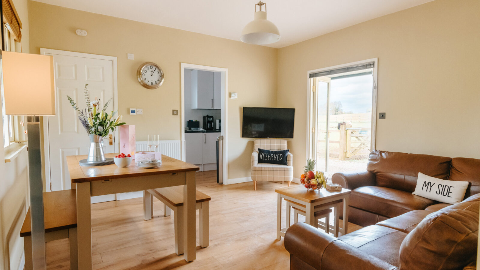 Cozy living room with a brown leather sofa, dining table, wall-mounted TV, and charming decor items reminiscent of a lodge. Sliding door leads outside.