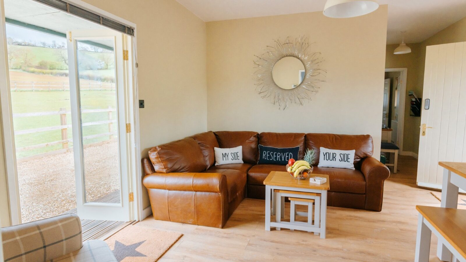 Cozy living room in the Chicken Shed Lodge, featuring a brown leather corner sofa, decorative pillows, and a round mirror. Door opens to a scenic view.