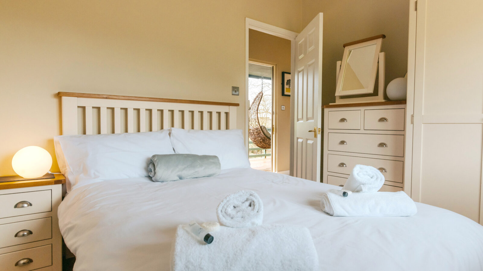 A tidy bedroom at the Chicken Shed Lodge, featuring a neatly made bed with white linens, two rolled towels, and a view into the sunlit adjoining room.