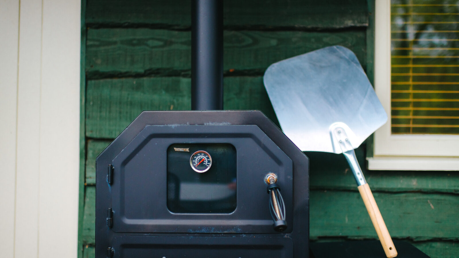 A black outdoor pizza oven with a temperature gauge stands next to a metal pizza peel, perfectly complementing the rustic charm of Chicken Shed Lodge against the green wooden wall.