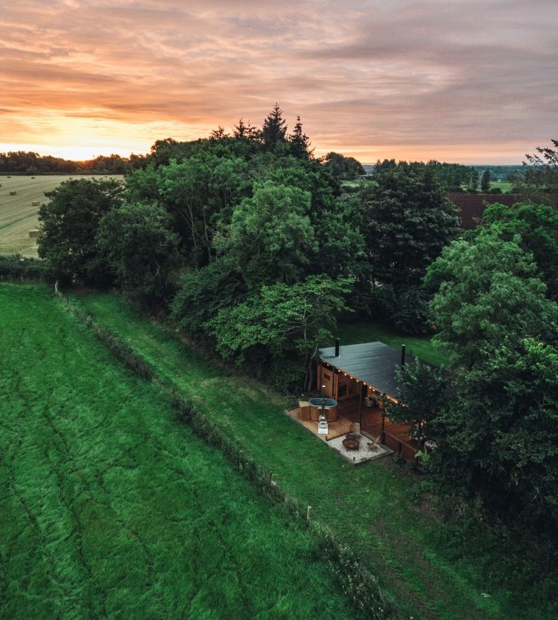 Aerial view of a cabin surrounded by trees at sunset, adjacent to a field, under the cloudy sky of Woodland Chase.