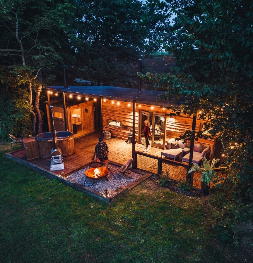 At Woodland Chase Cabins, a small cabin adorned with string lights features a hot tub and fire pit on a wooden deck nestled among the trees, where two people can be seen enjoying the serene forest setting.