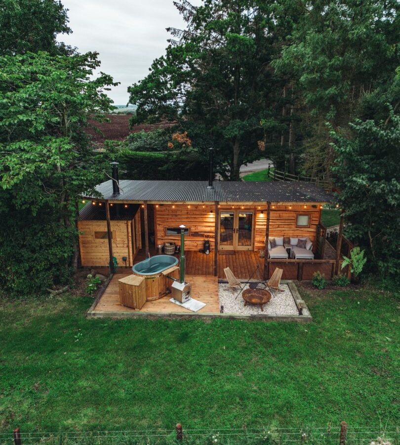 Aerial view of a woodland cabin featuring a hot tub, patio, and fire pit, nestled among trees and greenery.