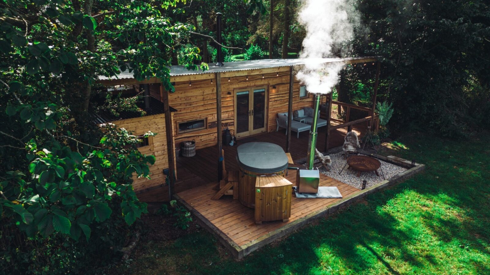 A charming wooden cabin nestled in Woodland Chase features a hot tub on its deck, surrounded by trees. On this sunny day, smoke gently rises from the chimney, adding to the serene atmosphere.